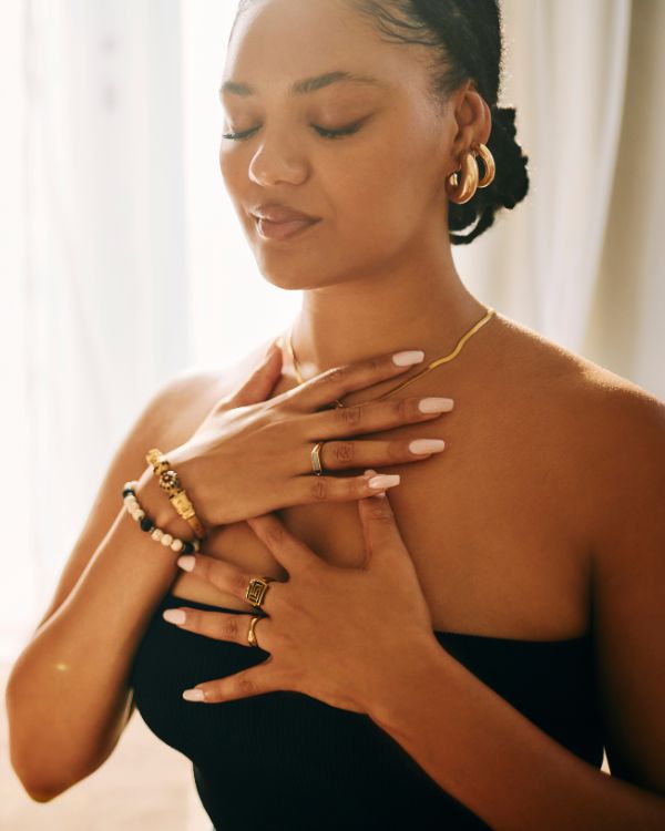 Home Page Woman practicing heart-centered meditation with eyes closed, wearing gold jewelry and healing crystal bracelets, in a softly lit wellness studio.